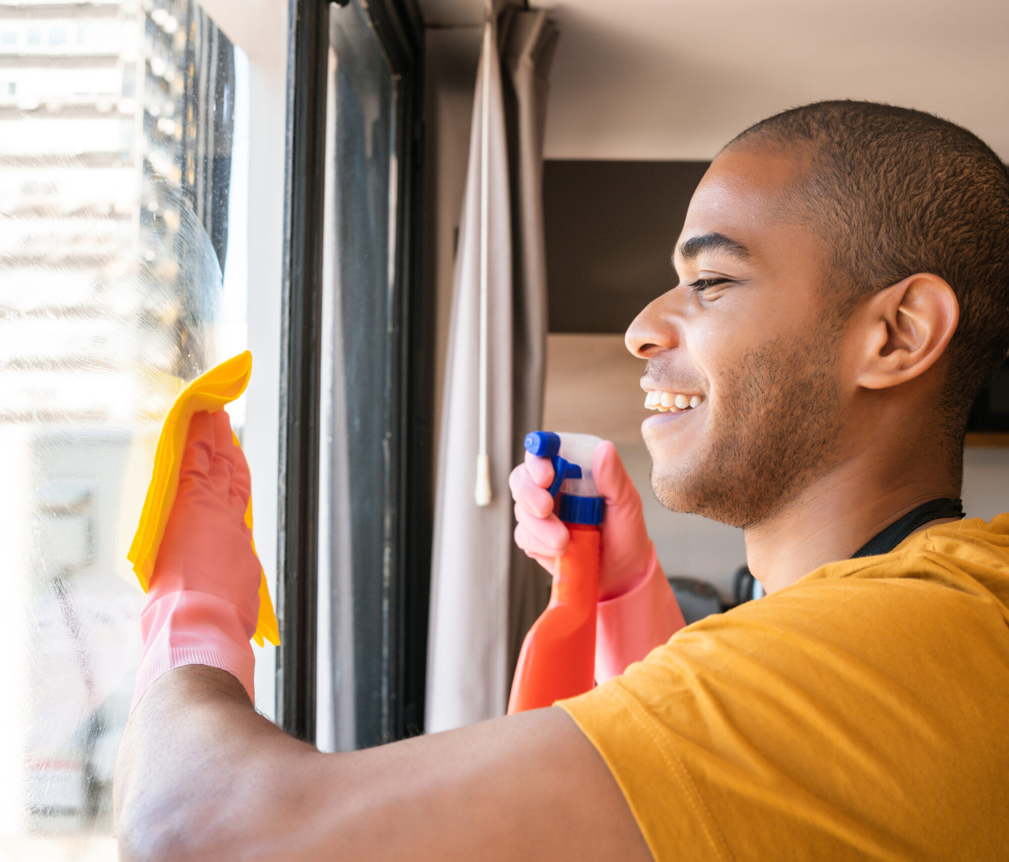 Portrait of young male housekeeper cleaning glass window at home. Housekeeping and cleaning concept.